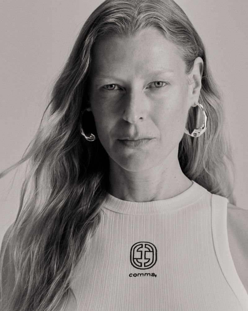 Headshot of a mature woman with long wavy hair, wearing hoop earrings and a white top with a logo against a light backdrop.