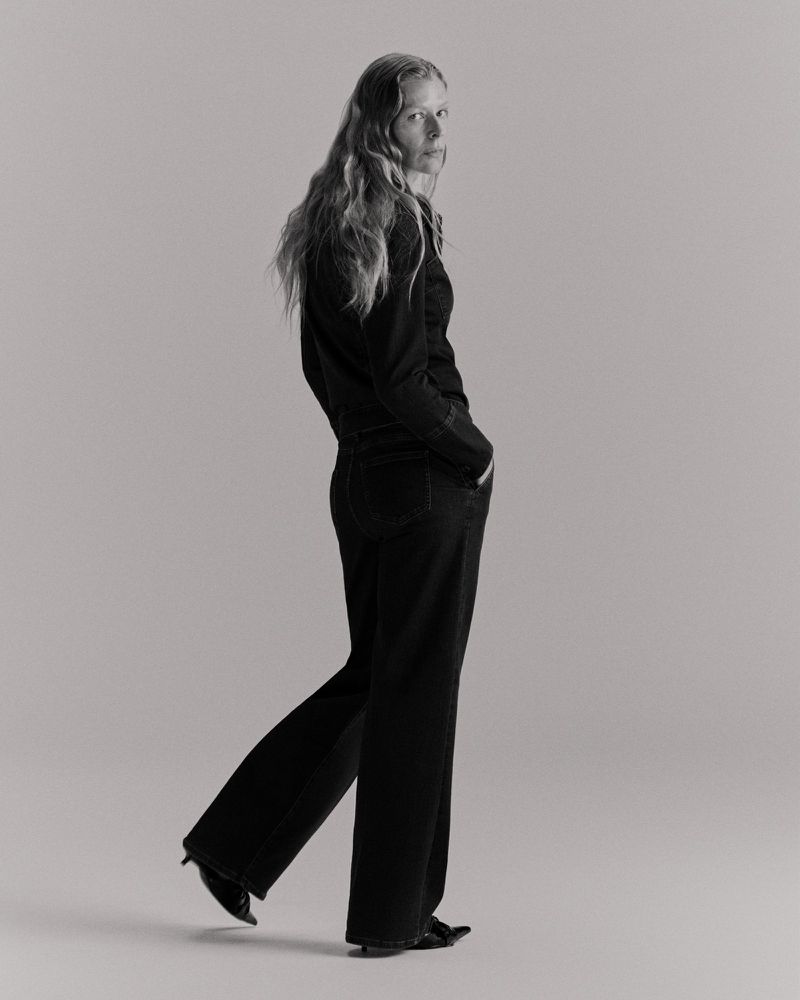 Woman with long wavy hair posing in dark wide-leg jeans and a black top, standing indoors in a photo studio against a plain background.