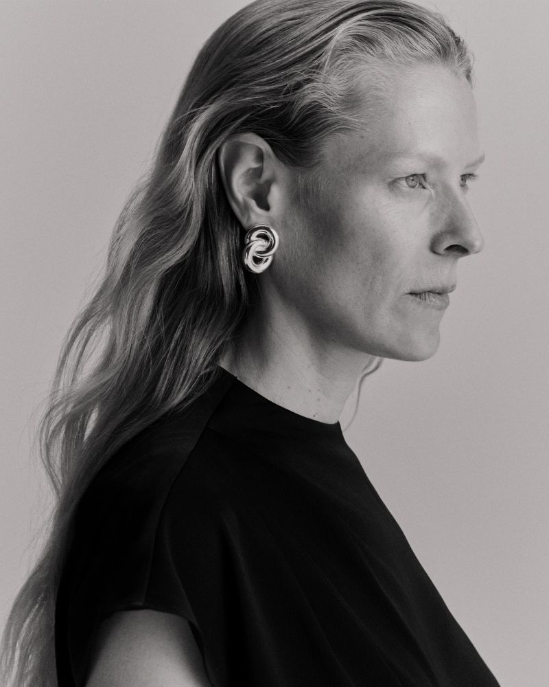 Profile portrait of a woman wearing a black top and silver sculptural earrings against a pale grey background