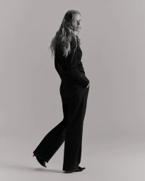 Model standing in a dark jumpsuit looking back over her shoulder against a neutral grey studio background.