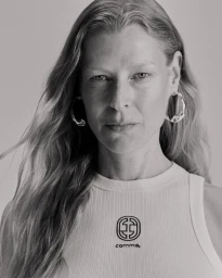 Portrait of a woman wearing a white ribbed tank top with a logo and large silver hoop earrings against a grey studio background.