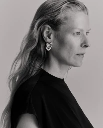 Profile portrait of a woman wearing a black top and silver sculptural earrings against a pale grey background