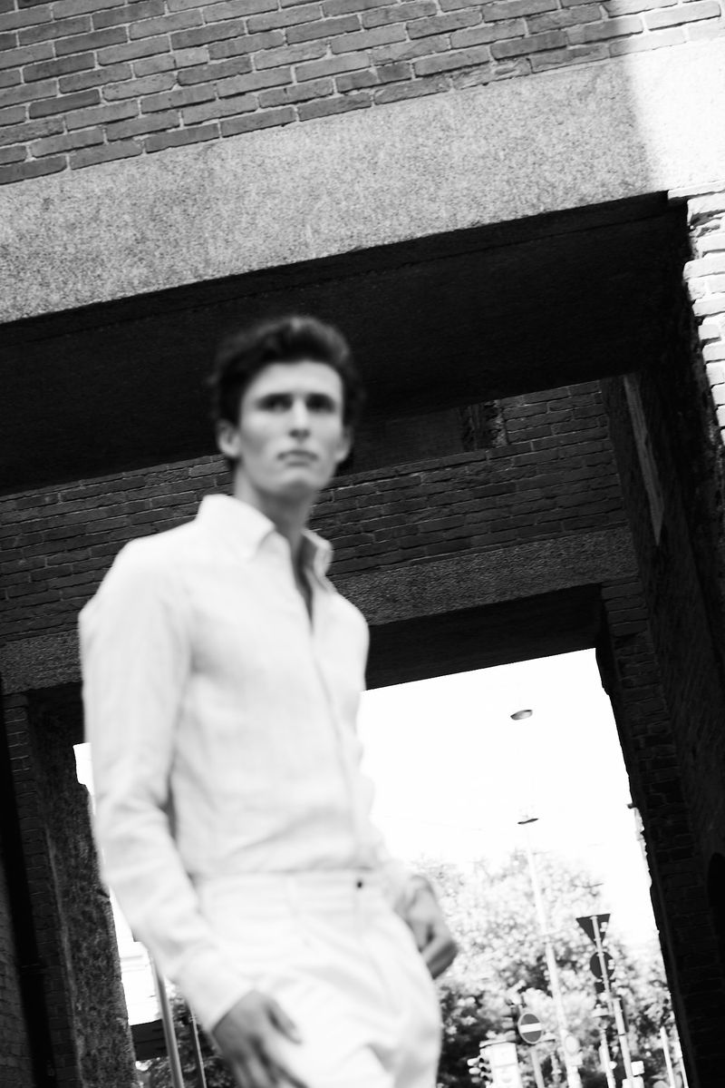 Man wearing a white shirt and trousers standing outdoors beneath a brick archway in a black and white photograph.