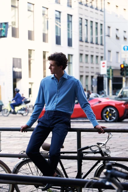 Man in blue shirt leans on railing with bicycles in an urban setting, with a red sports car visible in the background on a sunny day.