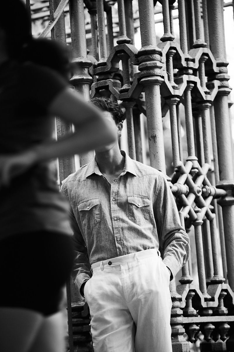 Black and white portrait of a man in button-down shirt and white pants leaning against an iron gate with another figure in the foreground.
