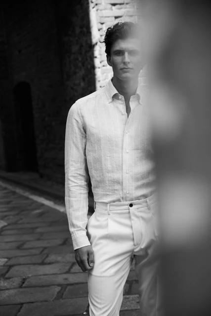 Monochrome portrait of a young adult man in a casual linen shirt and white pants standing on a cobblestone street.