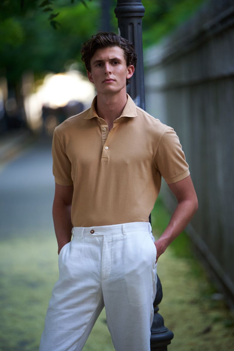 Outdoor portrait of a young adult male in a tan polo shirt and white pants, leaning against a lamp post in a blurred urban setting.