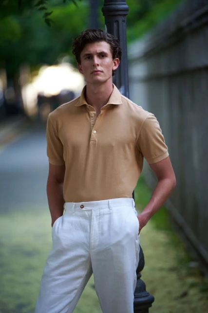 Outdoor portrait of a young adult male in a tan polo shirt and white pants, leaning against a lamp post in a blurred urban setting.