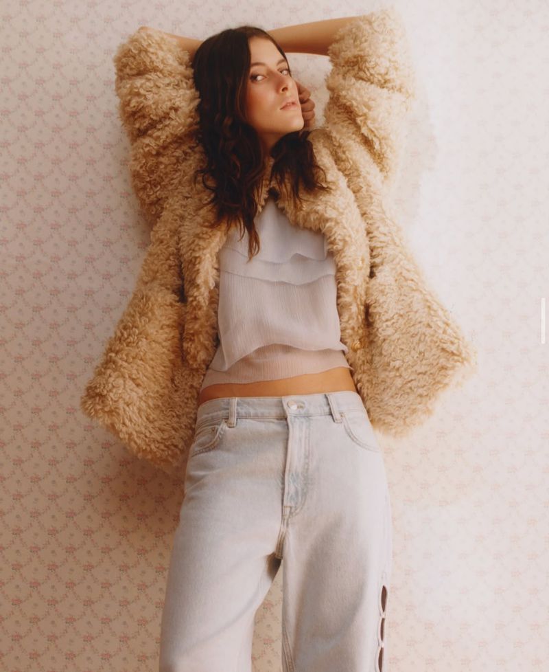 A young adult woman poses in a photo studio wearing a fuzzy jacket, crop top, and jeans against a patterned pink wallpaper backdrop.