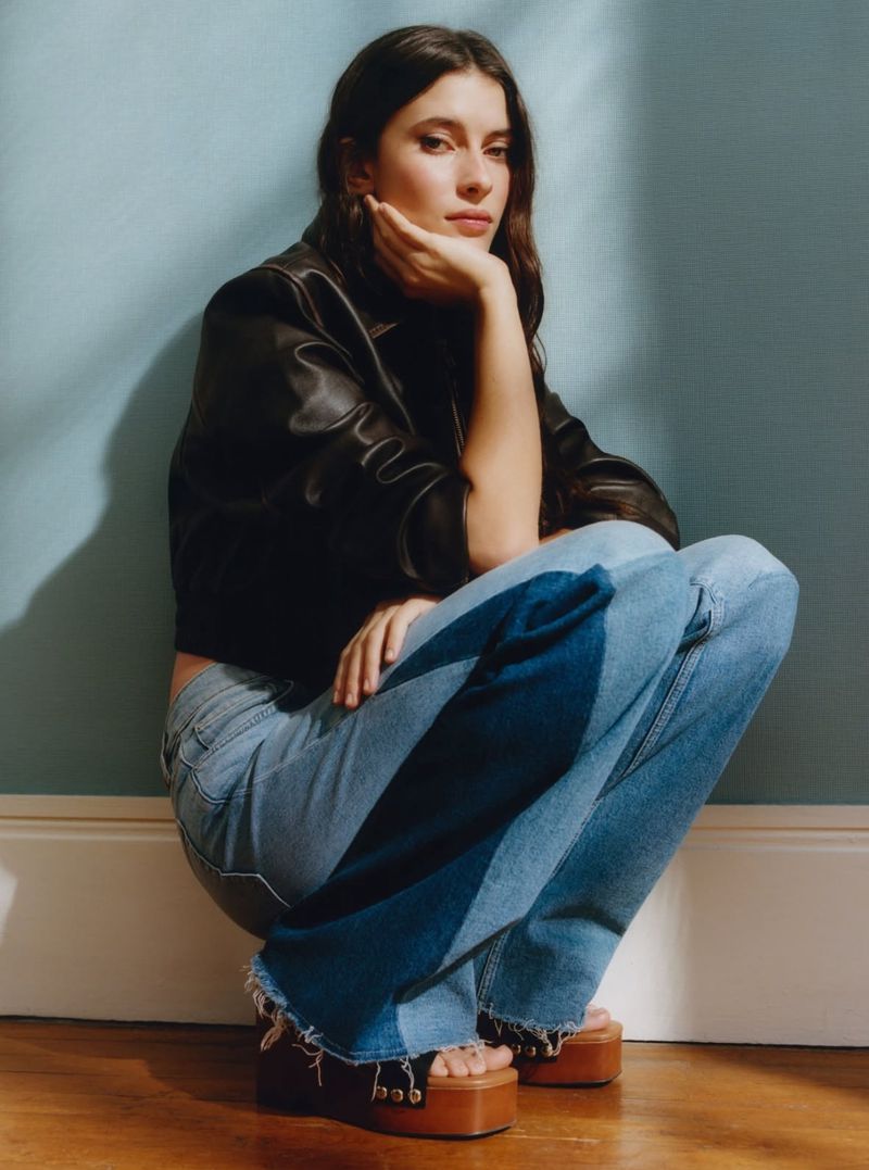 Young woman in stylish denim jeans and black leather jacket sits and poses in a studio with a light blue wall and wood flooring.