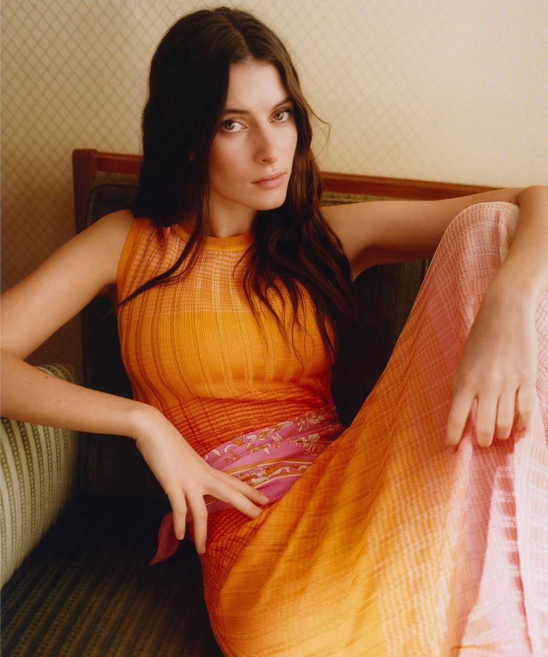 Elegant woman in orange top and patterned pants poses gracefully indoors, seated on a vintage couch, showcasing a blend of fashion and style.