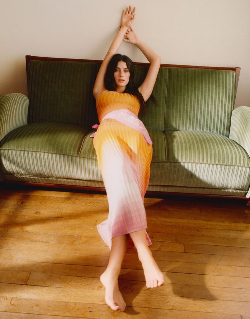 A young woman poses barefoot on a wooden floor, wearing a pastel orange-pink strapless dress, next to a green couch in a studio setting.