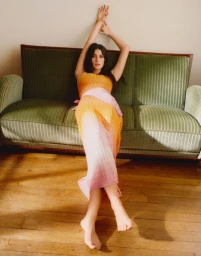 Model lying on a wooden floor leaning against a green striped sofa wearing an orange and pink gradient dress