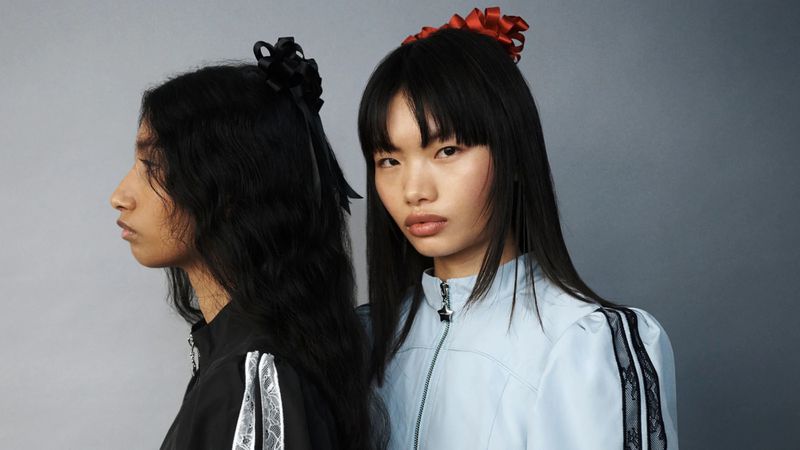 Two Asian women pose in coordinating black and blue track jackets with hair bows against a gray background in a studio-lit setting.