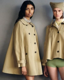Two female models wearing tan trench coats and a head covering standing against a grey seamless studio background.
