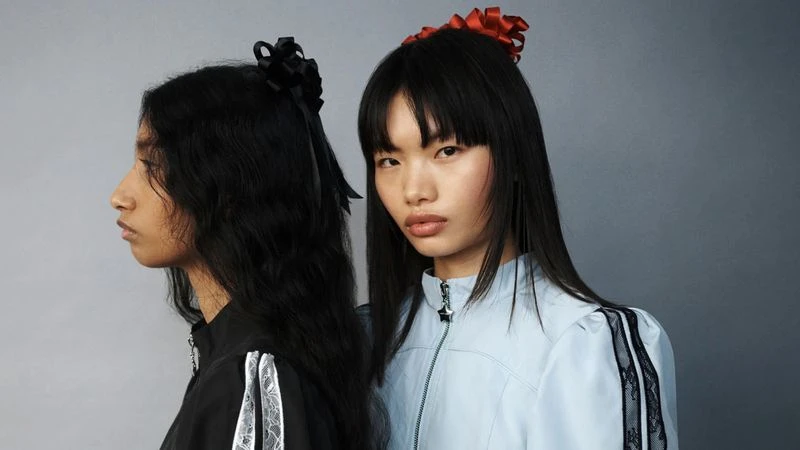 Two female models posing in profile and front view against a neutral grey studio backdrop for Sandy Liang Spring/Summer 2025 campaign.