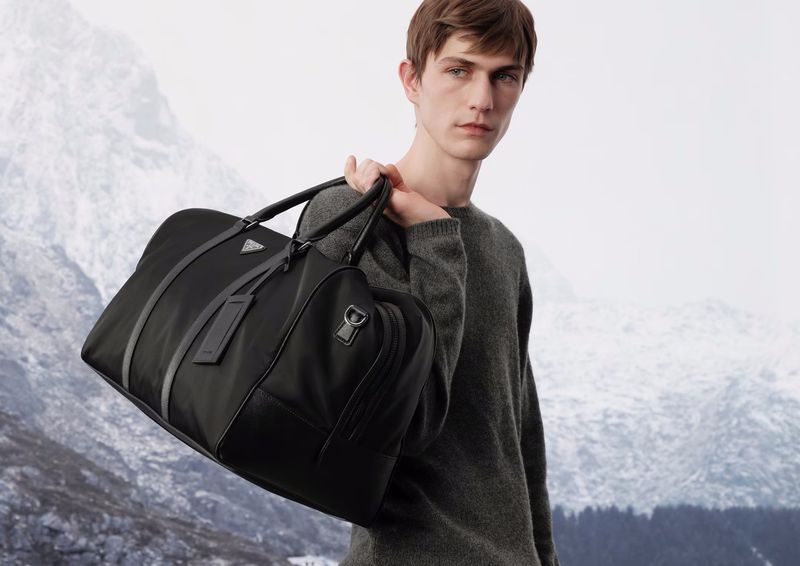 A stylish black Prada duffel bag is carried by a slender man with light brown hair wearing a grey sweater, against a mountain backdrop.