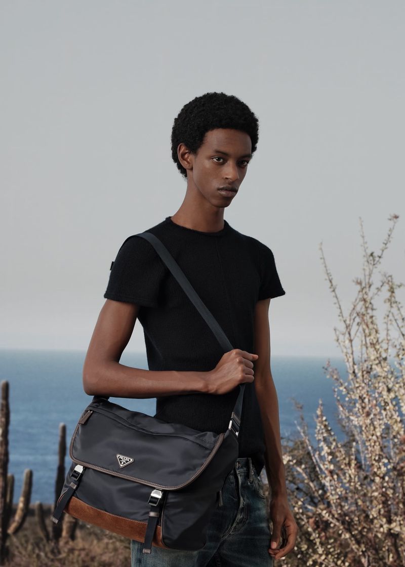 An adult male model poses outdoors wearing a black t-shirt and jeans, carrying a Prada messenger bag, with a sky and ocean background.