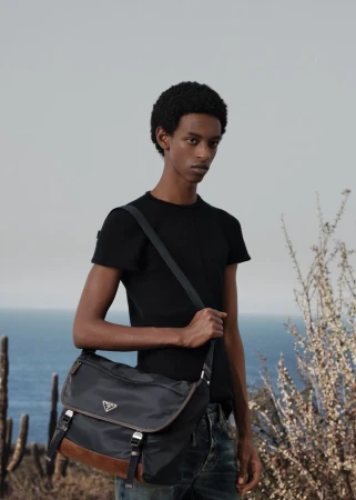 An adult male model poses outdoors wearing a black t-shirt and jeans, carrying a Prada messenger bag, with a sky and ocean background.