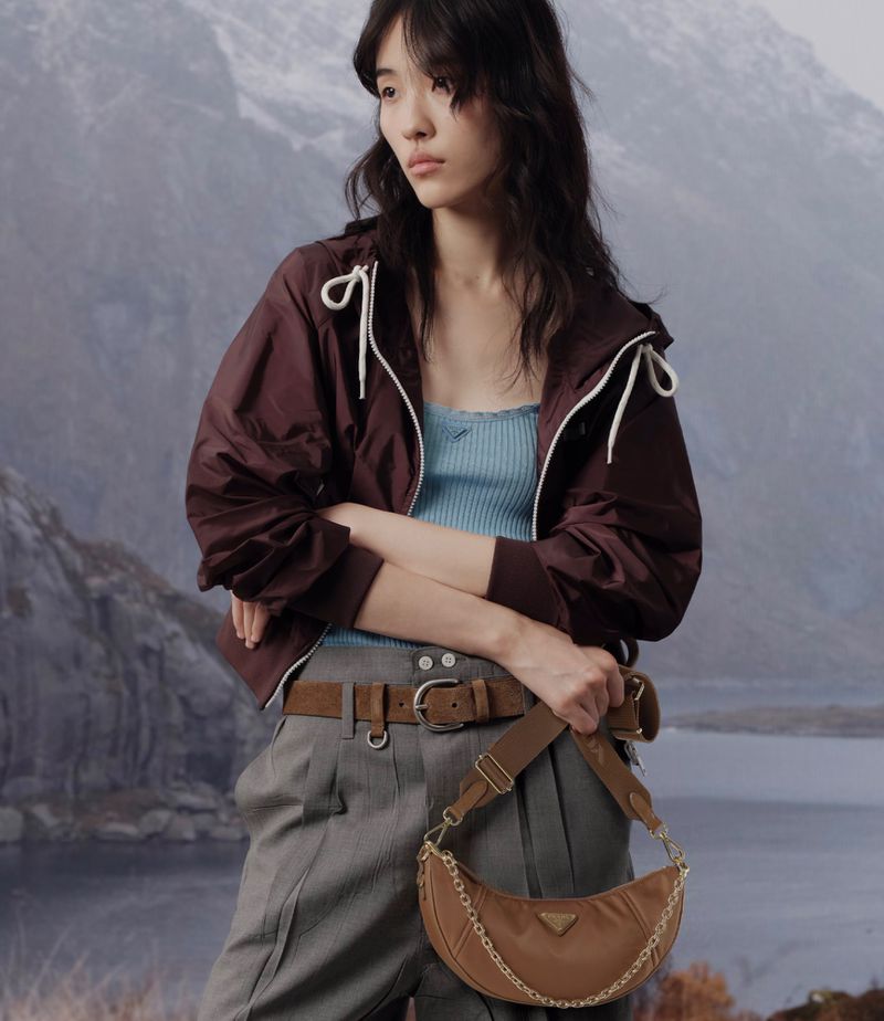 A fashionable woman poses in front of a mountain range, wearing a windbreaker, pants, and a brown purse with a gold chain.
