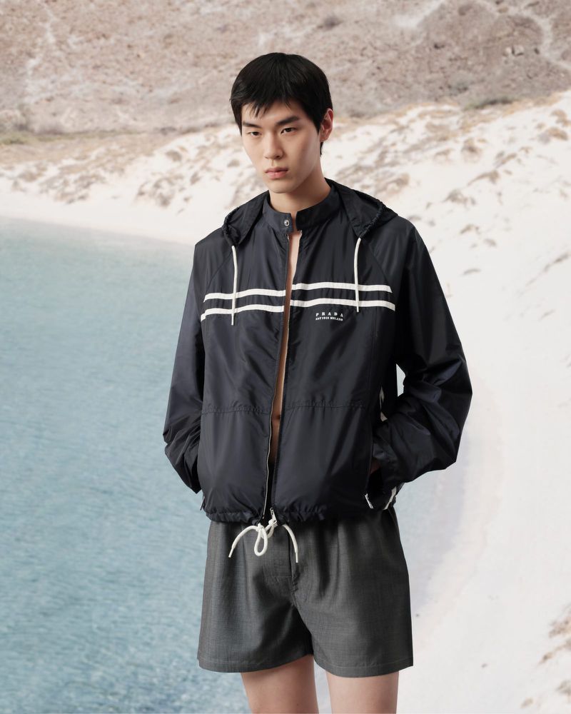 Asian male fashion model poses outdoors on a beach wearing a striped hooded windbreaker and shorts in a medium shot.
