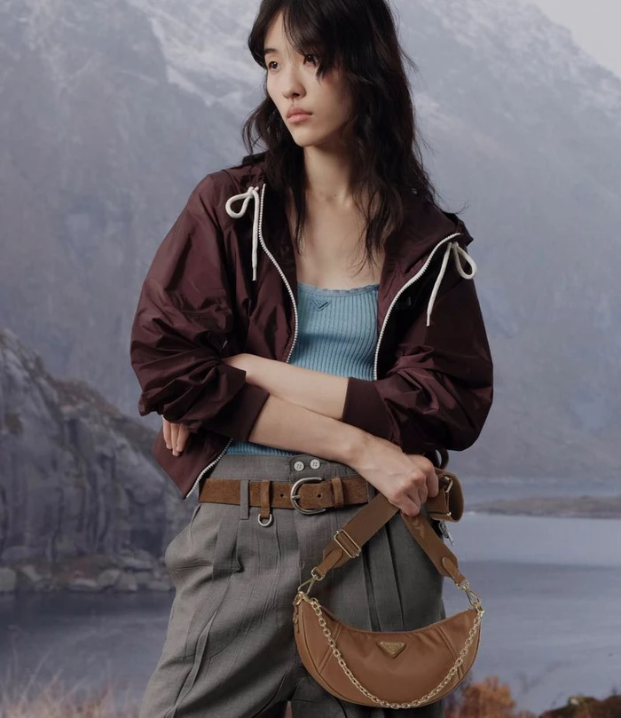 Model wearing a dark maroon jacket and grey trousers holding a brown shoulder bag against a mountain landscape background