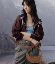 Model wearing a dark maroon jacket and grey trousers holding a brown shoulder bag against a mountain landscape background
