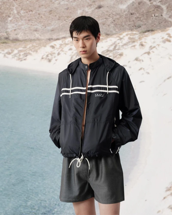 East Asian male model wearing Prada black nylon zip jacket and grey shorts standing near a sandy coastal beach.