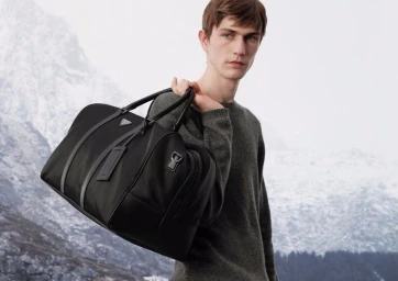 A male model holding a black travel bag over his shoulder against a snowy mountain landscape.