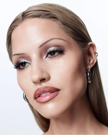 Close-up portrait of a woman with light blonde hair, silver eyeshadow, and glossy lips against a plain white background.