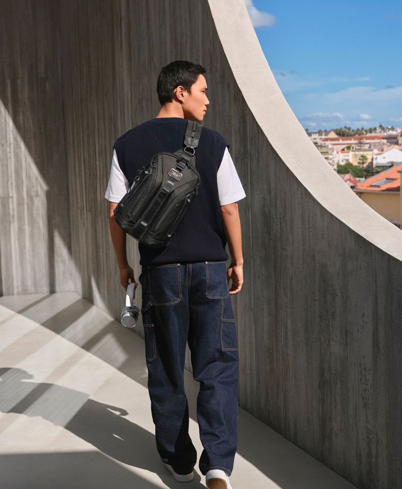 Man stands on a walkway wearing a sling bag, navy vest, white shirt, and jeans with a cityscape and blue sky background.