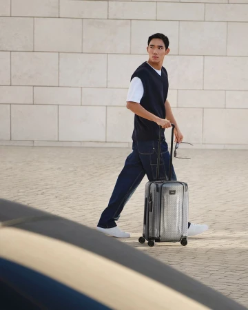 Model walking with a silver rolling suitcase in front of a stone wall for the Tumi 50th Anniversary campaign