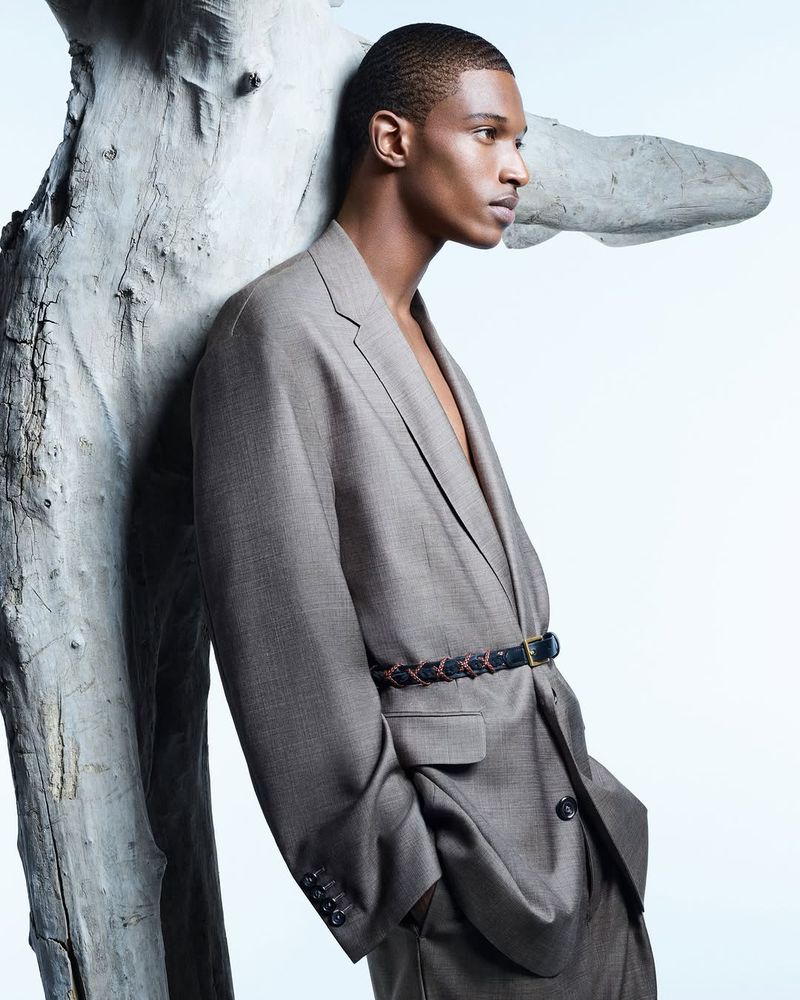 Portrait of African-American male fashion model in an oversized grey suit leaning against a textured tree branch in a photo studio.