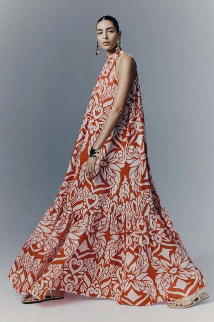 A woman in a red and white print sleeveless dress and sandals poses in a studio, showcasing a modern fashion look.