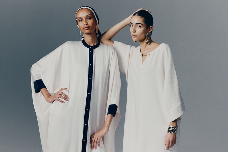 Two women in white dresses pose in a studio. One wears a headscarf with black trim, and the other wears gold jewelry. Fashion shot.