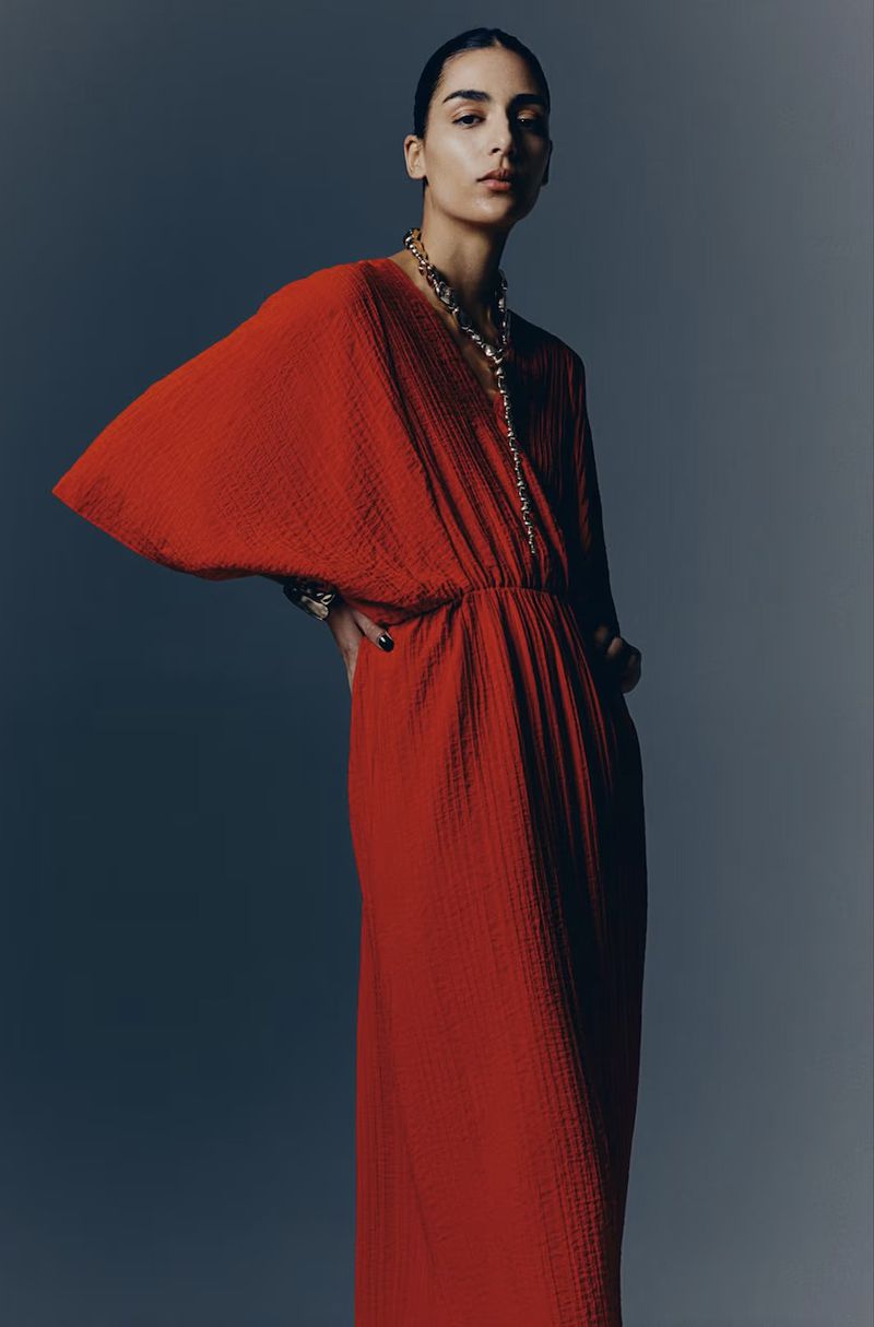 Model wearing a vibrant red pleated dress and a gold necklace against a grey studio backdrop for H&M Limited Edition 2025.