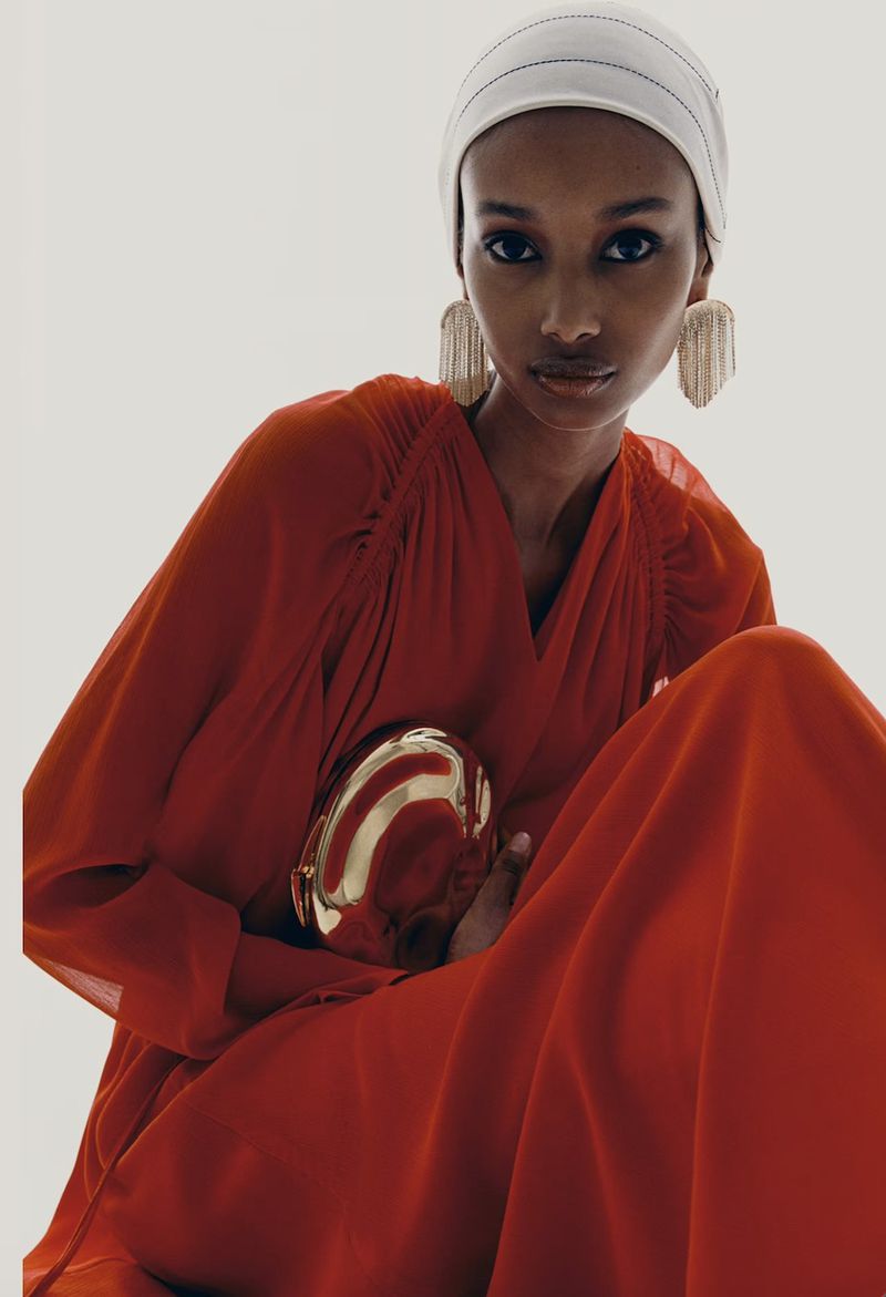 Woman in red dress with head covering and metallic purse poses in photo studio with bright lighting for fashion editorial shoot.