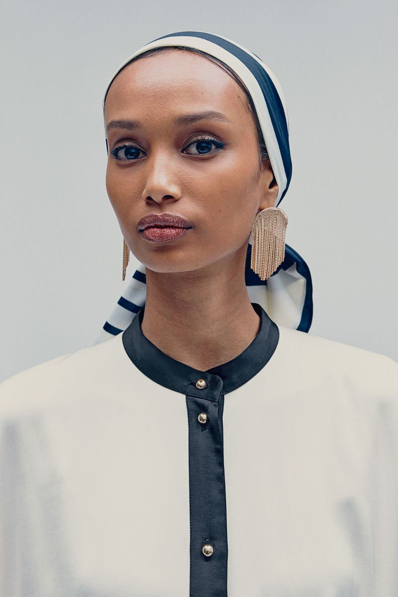 Close-up portrait of an adult female model wearing a striped headscarf, long gold fringe earrings, and a contrasting dress.