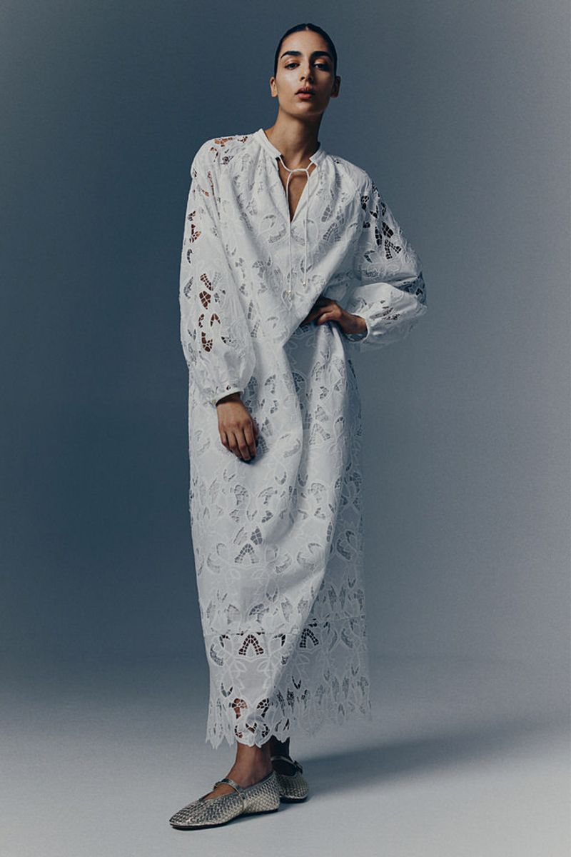 A fashionable adult woman wearing a white, long-sleeved, eyelet-detailed dress and flat shoes poses against a gradient gray background.