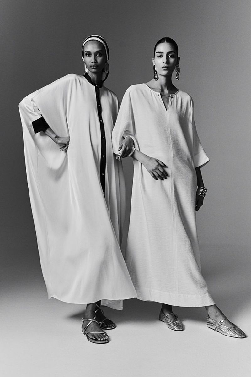 Two adult women pose in flowing white dresses in a studio shot. One woman wears a headscarf. Both models wear metallic flats and jewelry.