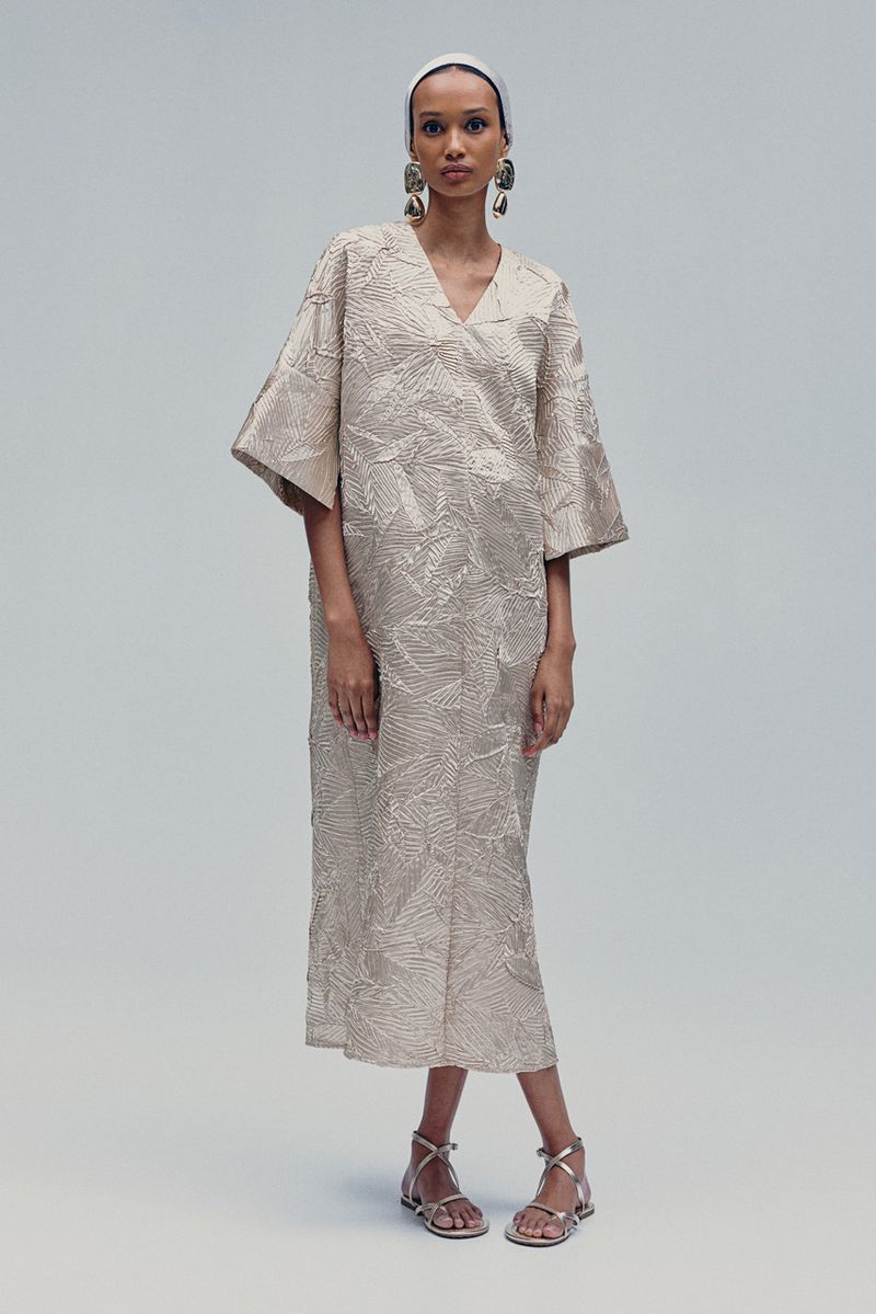 A slender woman poses in a studio, showcasing a beige leaf-patterned midi dress with wide sleeves and silver sandals, complemented by large earrings.