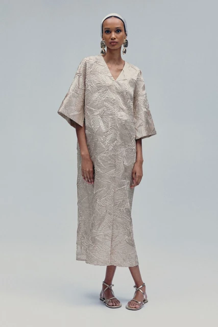 A slender woman poses in a studio, showcasing a beige leaf-patterned midi dress with wide sleeves and silver sandals, complemented by large earrings.