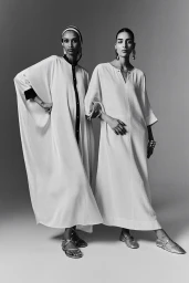 Two female models wearing flowing white dresses posing together against a plain studio background.