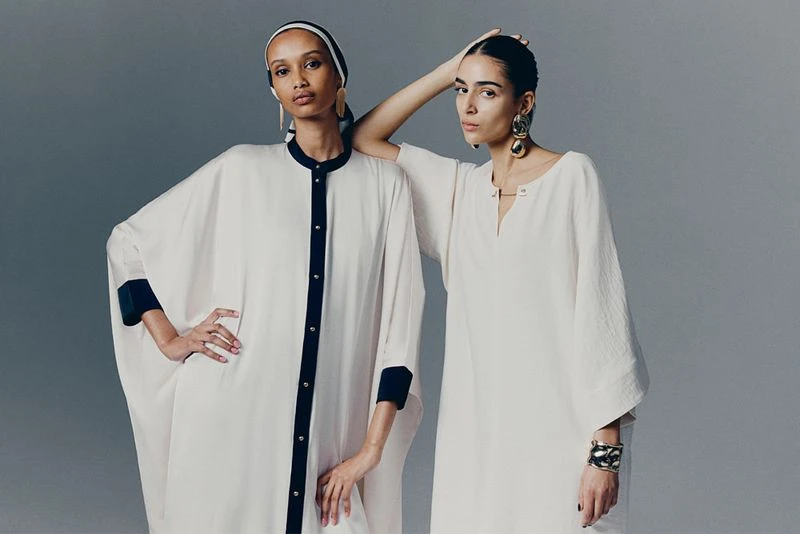 Two female models wearing white kaftans with black trim against a neutral grey studio background for H&M.