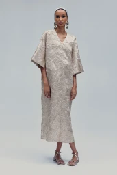 Model Nora Attal wearing a textured cream dress and silver sandals against a grey seamless background