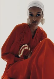 Model in red dress wearing a white headscarf and holding a gold clutch against white background