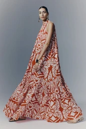 Model wearing a flowing red and white patterned halter maxi dress against a plain grey studio background.