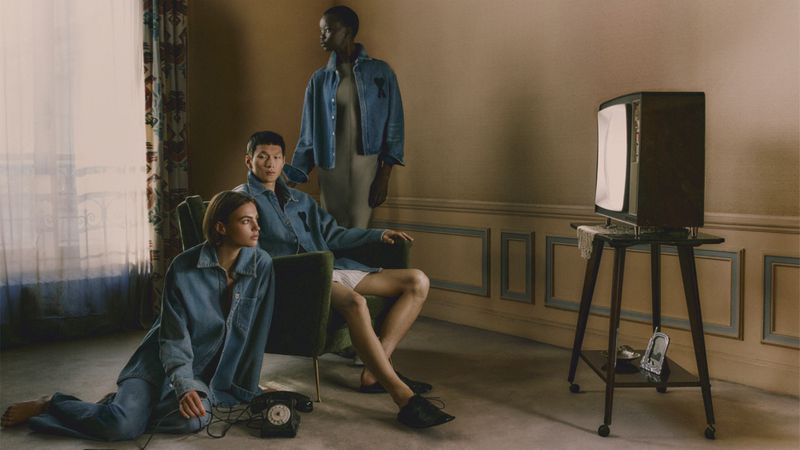 Three individuals in denim attire gather indoors around a vintage television set in a room featuring floral curtains and period decor.