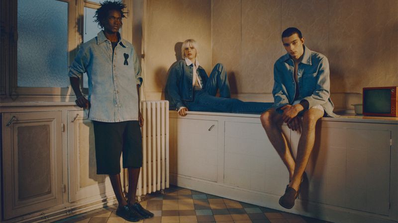 Three fashion models pose indoors against a wall in denim and casual attire, illuminated by natural light, conveying a relaxed and stylish aesthetic.