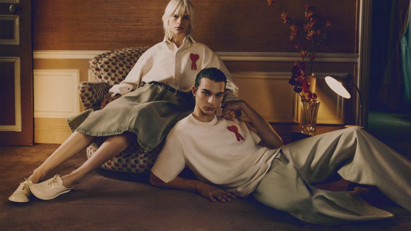 Two fashion models pose in a warm-toned interior: a seated blonde woman and a dark-haired man reclining on the floor, both wearing AMI apparel.